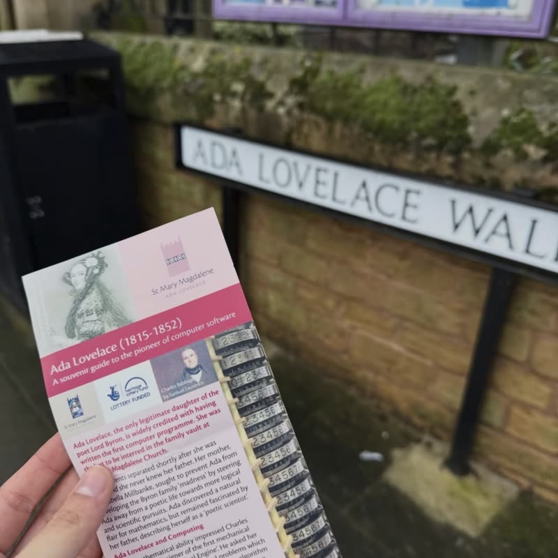 A souvenir leaflet about Ada Lovelace held up in front of the Ada Lovelace Walk street sign.