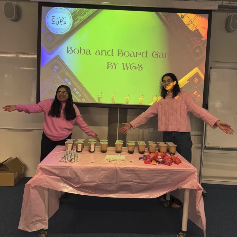 Two committee members with their arms out in front of a table of boba cups, under a projector screen saying Boba and Board Games by WCS.