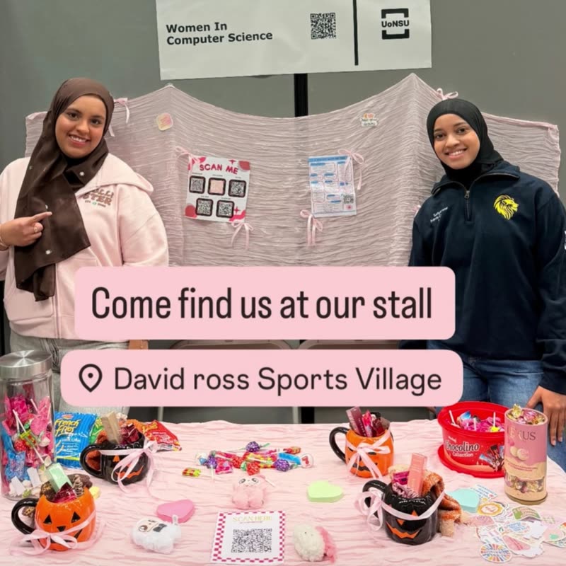 Two committee members smiling at the WCS stall in David Ross Sports Village, with sweets and pumpkin mugs on a pink table.