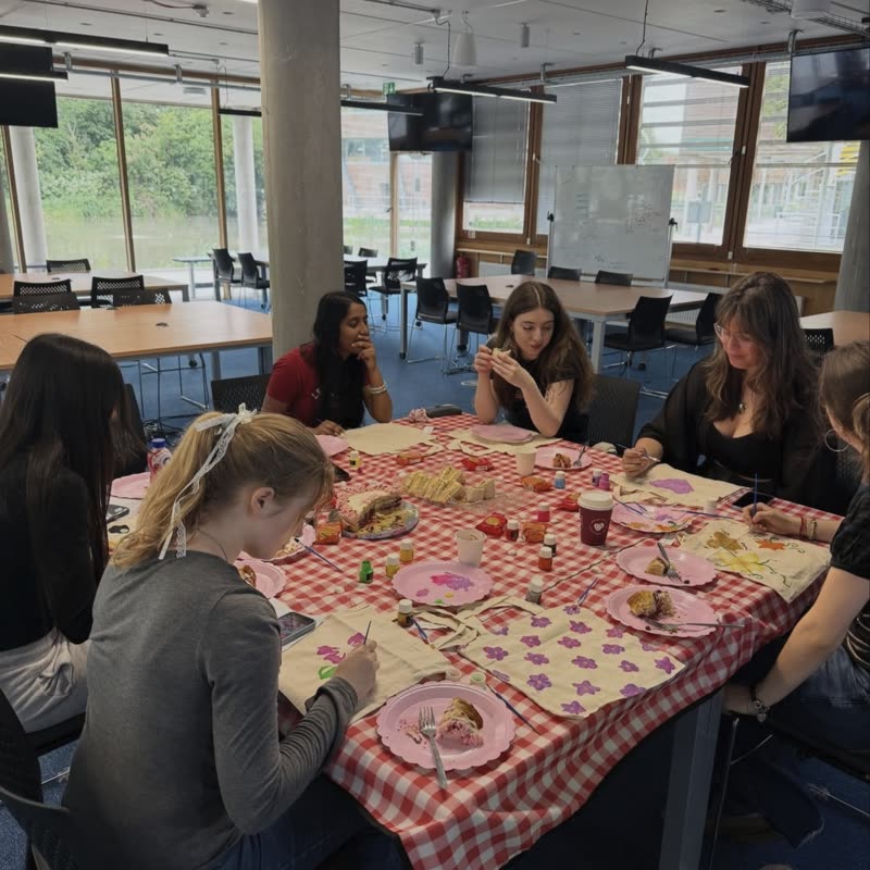 Members painting tote bags around a table with a red checked cloth, pink plates and picnic food.