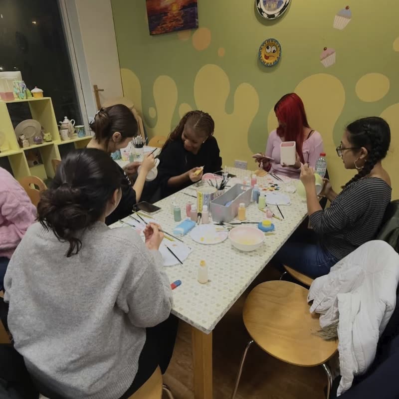 Members painting mugs and bowls around a table in a pottery cafe.