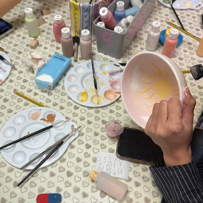 A hand holding a pink bowl with a yellow sun painted inside, surrounded by paint palettes and brushes.